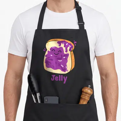 Jelly couple butter apron - Cartoon tan toast with smiling purple jelly face and purple Jelly text — shown on a black cotton canvas apron against a white background