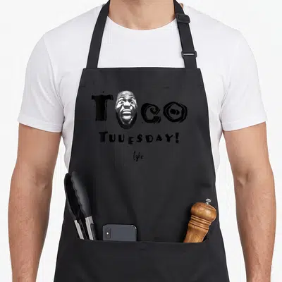 Los taco apron - Black and white graphic of a screaming man's face replacing the O in TCO with TUUESDAY text below — shown on a black cotton canvas apron against a white background