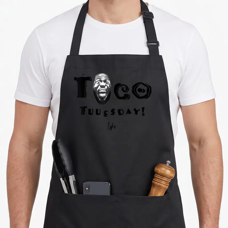 Los taco apron - Black and white graphic of a screaming man's face replacing the O in TCO with TUUESDAY text below — shown on a black cotton canvas apron against a white background