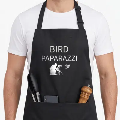 Birdwatching bird aprons - Distressed white BIRD PAPARAZZI text above kneeling photographer and flying bird silhouette — shown on a black cotton canvas apron against a white background