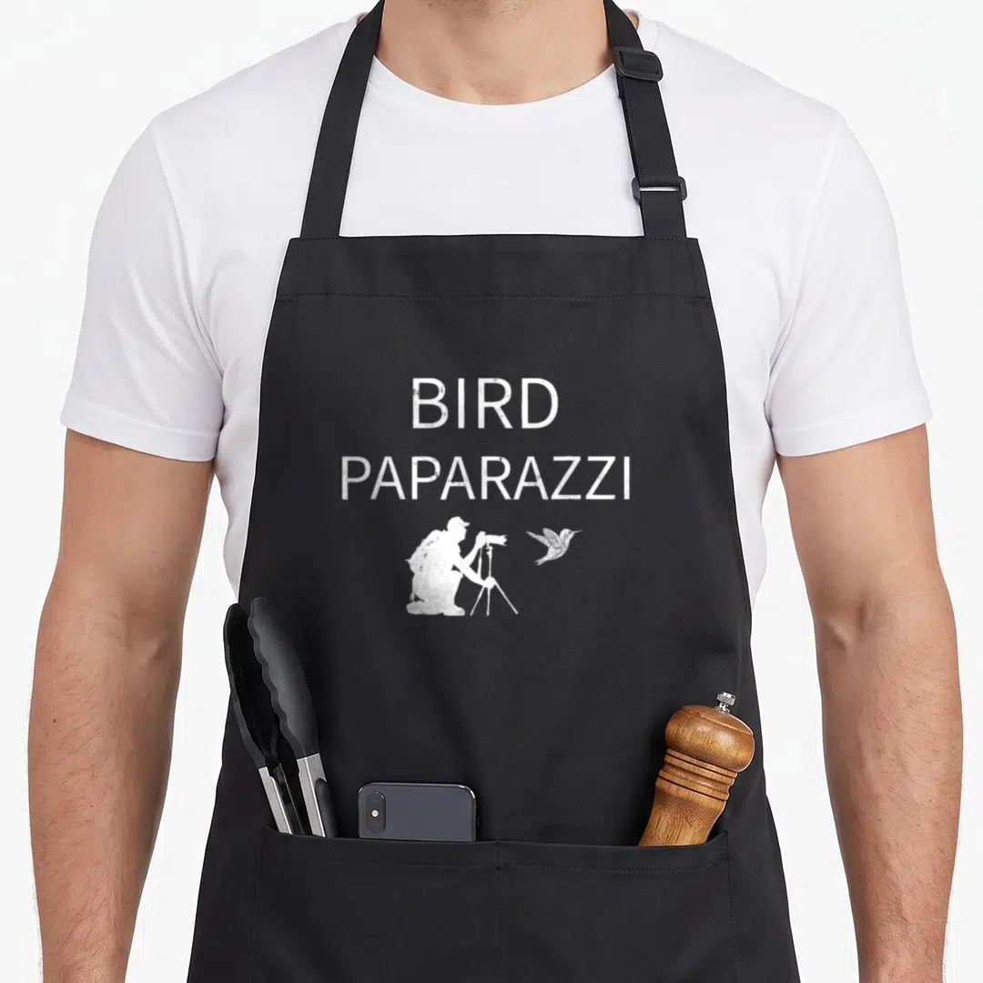 Birdwatching bird aprons - Distressed white BIRD PAPARAZZI text above kneeling photographer and flying bird silhouette — shown on a black cotton canvas apron against a white background