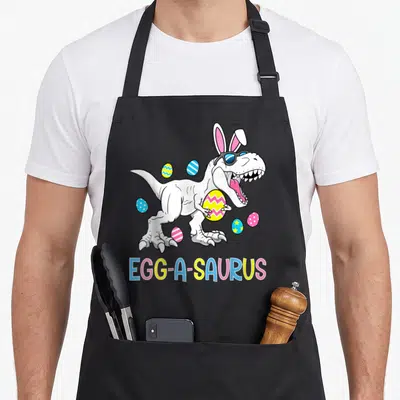 Saurus easter egg apron - White dinosaur with bunny ears holding egg, text EGG-A-SAURUS — shown on a black cotton canvas apron against a white background