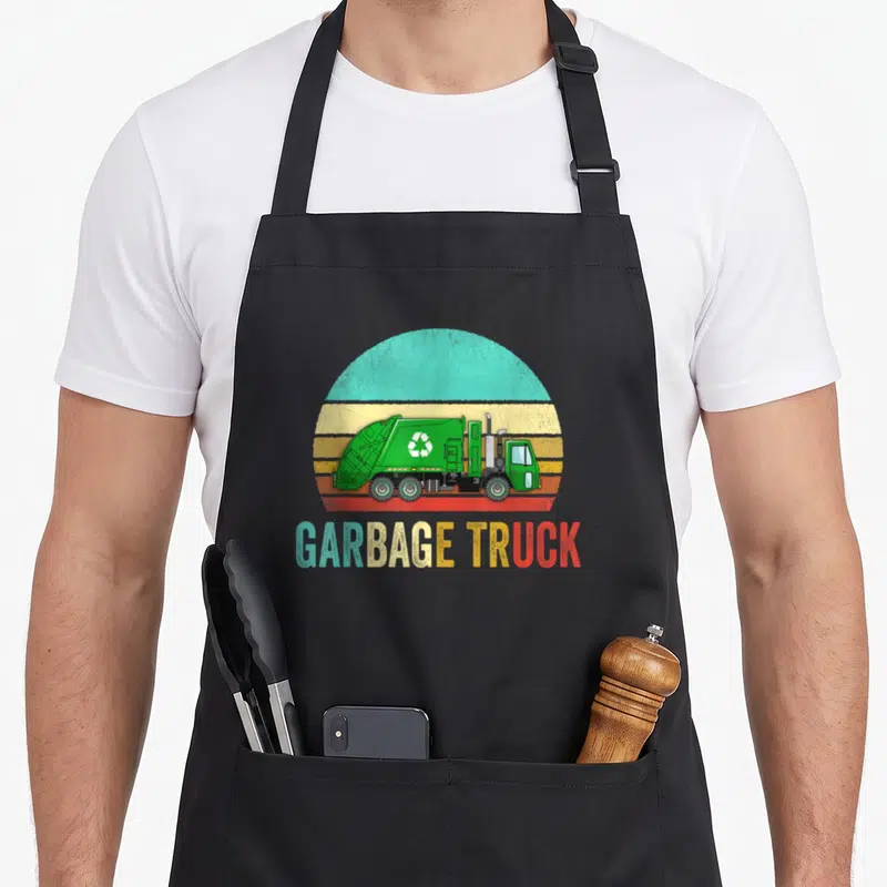 Garbage sunset truck apron - Green garbage truck illustration in front of a striped retro sun with gradient text below — shown on a black cotton canvas apron against a white background