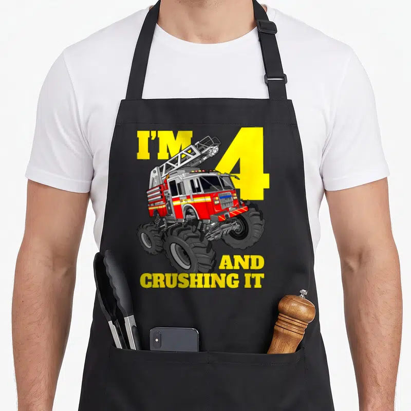Four firefighter truck apron - Cartoon monster fire truck over yellow number 4 with text I'M 4 AND CRUSHING IT — shown on a black cotton canvas apron against a white background