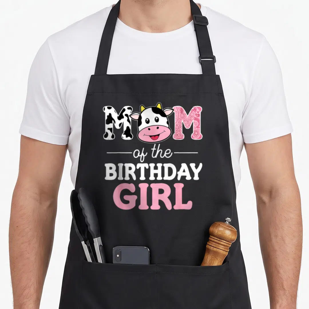 Birthday cow print apron - Cartoon cow head replaces O in MOM text with pink swirl and black spot patterns above — shown on a black cotton canvas apron against a white background