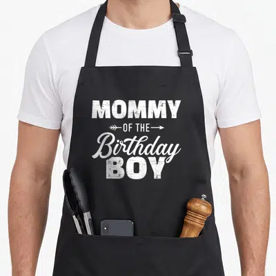Son family cooking aprons - White MOMMY OF THE Birthday BOY text with arrows on black background — shown on a black cotton canvas apron against a white background