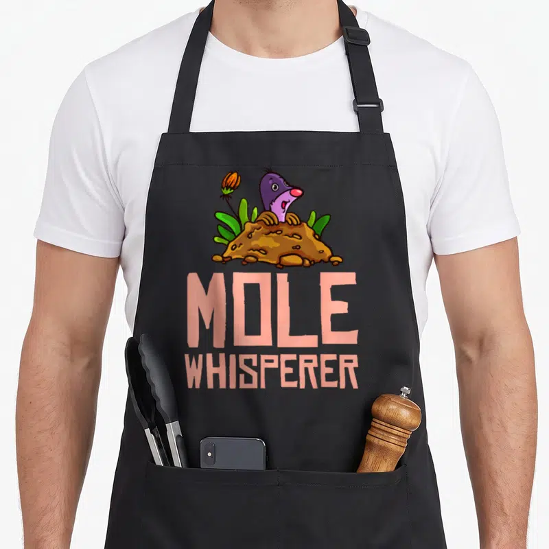 Mole hunter chemistry apron - Cartoon purple mole in brown dirt mound with pink stacked text reading MOLE WHISPERER — shown on a black cotton canvas apron against a white background