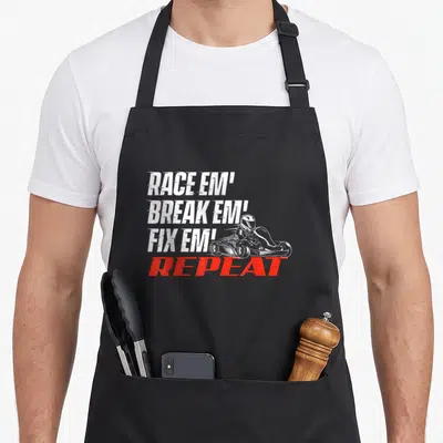 Break racing apron - Stacked RACE EM BREAK EM FIX EM REPEAT text with go kart driver line art illustration — shown on a black cotton canvas apron against a white background