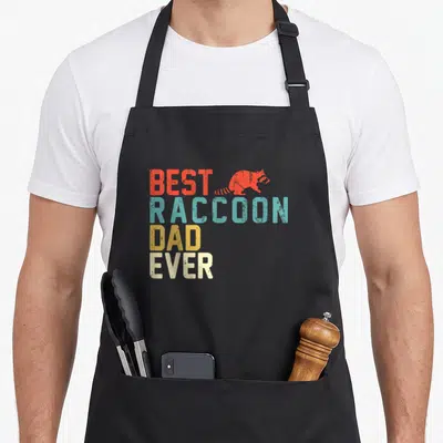 Funny raccoon dad apron - Stacked retro text BEST RACCOON DAD EVER in red teal yellow and off-white with raccoon — shown on a black cotton canvas apron against a white background