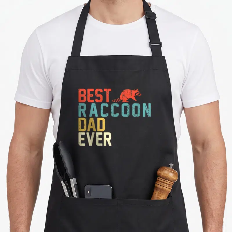 Funny raccoon dad apron - Stacked retro text BEST RACCOON DAD EVER in red teal yellow and off-white with raccoon — shown on a black cotton canvas apron against a white background