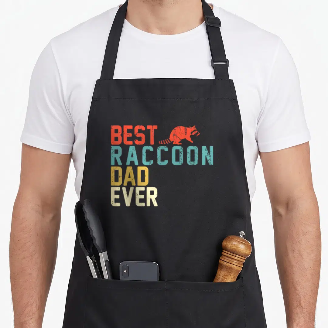 Funny raccoon dad apron - Stacked retro text BEST RACCOON DAD EVER in red teal yellow and off-white with raccoon — shown on a black cotton canvas apron against a white background