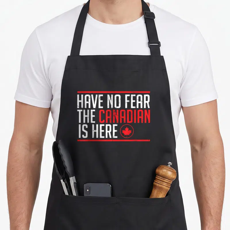 Fear canadian aprons - Distressed white and red text reading HAVE NO FEAR THE CANADIAN IS HERE with a maple leaf icon — shown on a black cotton canvas apron against a white background