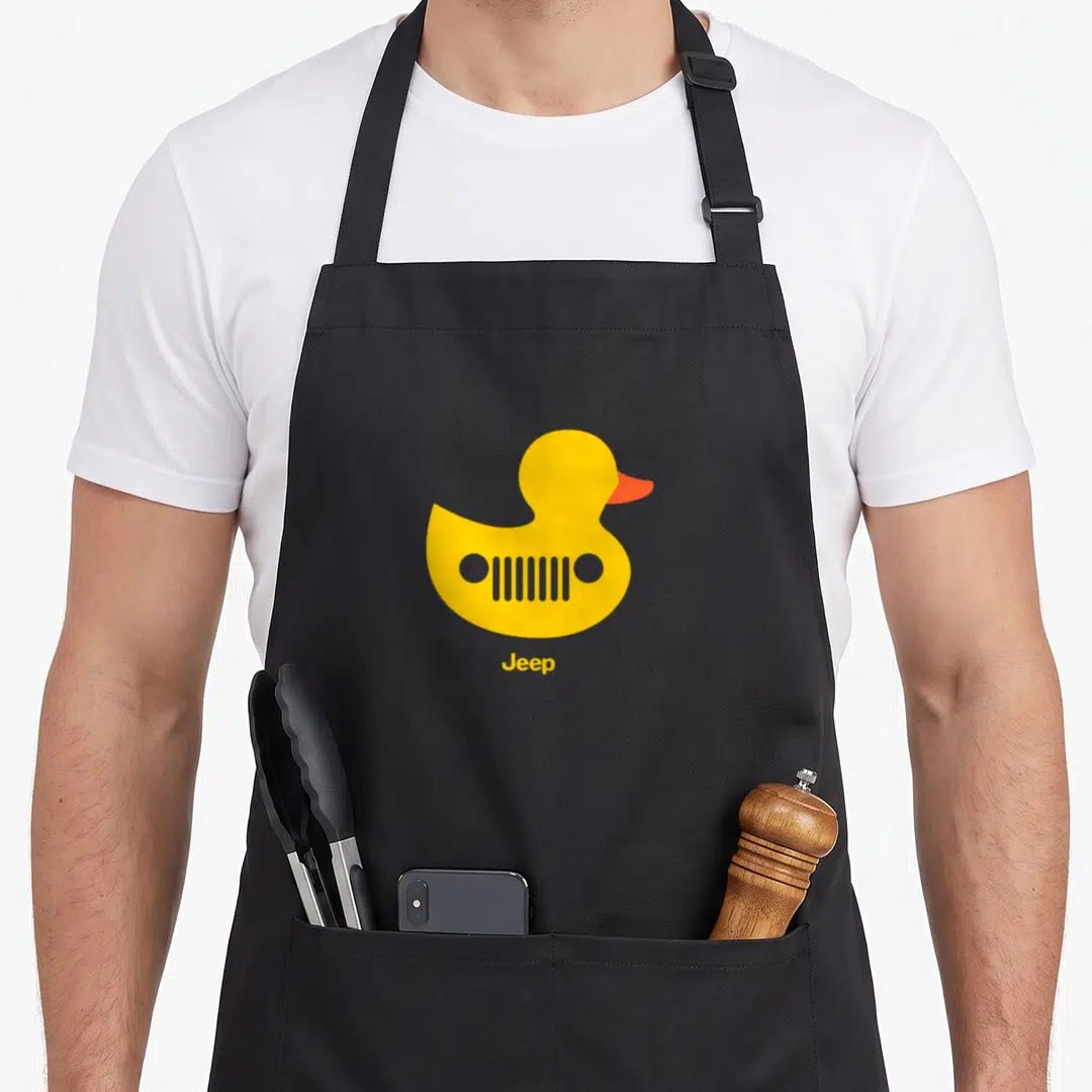 Jeep grille duck apron - Yellow rubber duck with car grille body and orange beak, text Jeep below — shown on a black cotton canvas apron against a white background