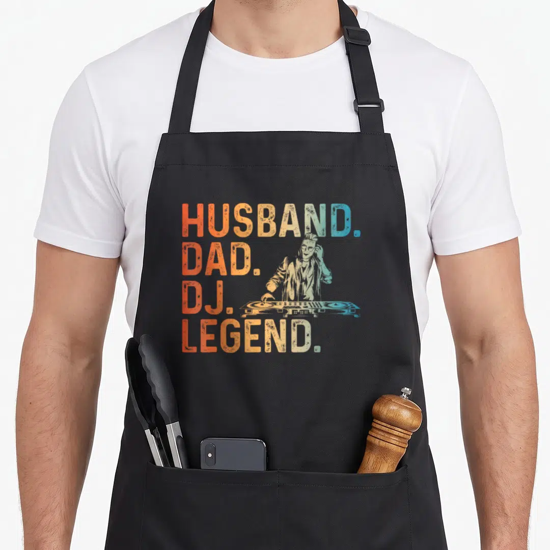 Producer husband apron - Stacked text Husband Dad DJ Legend with illustrated DJ and turntable in orange and blue — shown on a black cotton canvas apron against a white background