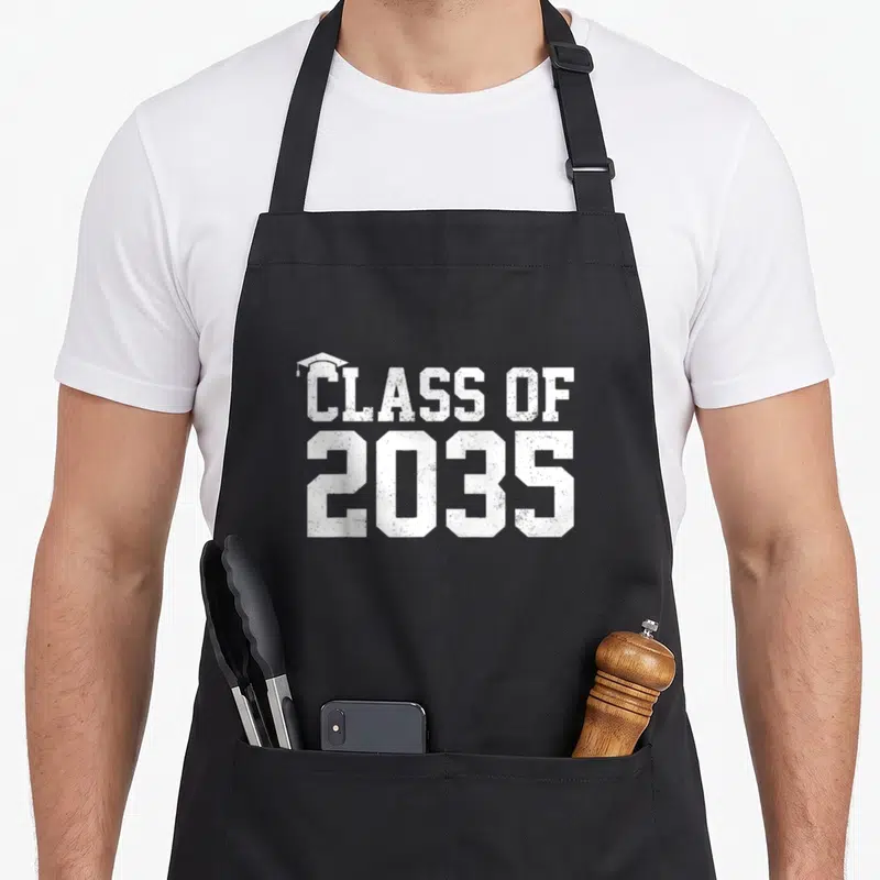 Grow class first apron - White CLASS OF 2035 text with graduation cap on top of C — shown on a black cotton canvas apron against a white background