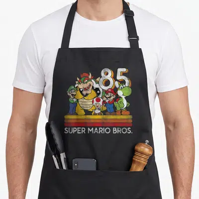 Character holiday apron - Retro cluster of characters with 85 and Super Mario Bros text above three horizontal stripes — shown on a black cotton canvas apron against a white background
