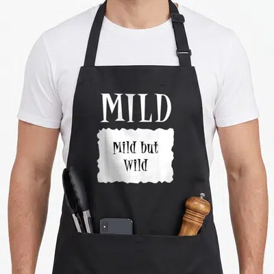 Mild taco apron - White MILD text above torn paper reading Mild but Wild in black gothic font — shown on a black cotton canvas apron against a white background