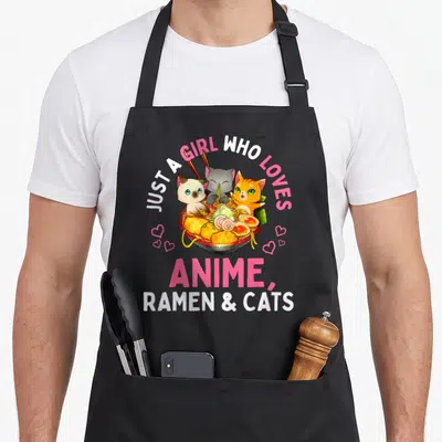 Manga cat apron - Three cartoon cats in a ramen bowl with pink hearts and text about loving anime ramen and cats — shown on a black cotton canvas apron against a white background