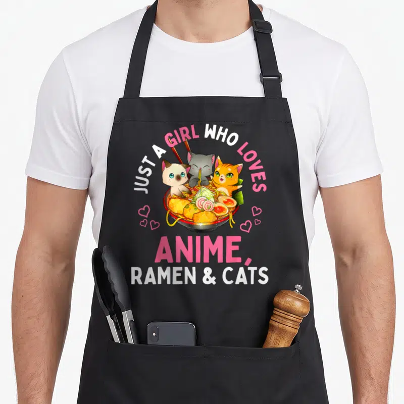 Manga cat apron - Three cartoon cats in a ramen bowl with pink hearts and text about loving anime ramen and cats — shown on a black cotton canvas apron against a white background