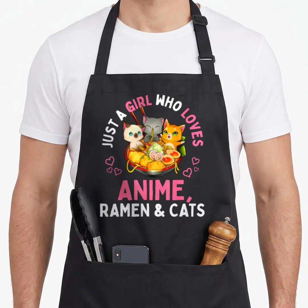 Manga cat apron - Three cartoon cats in a ramen bowl with pink hearts and text about loving anime ramen and cats — shown on a black cotton canvas apron against a white background
