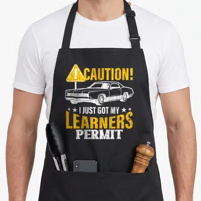 Learner car apron - Distressed yellow caution sign and white car with I just got my learners permit text — shown on a black cotton canvas apron against a white background