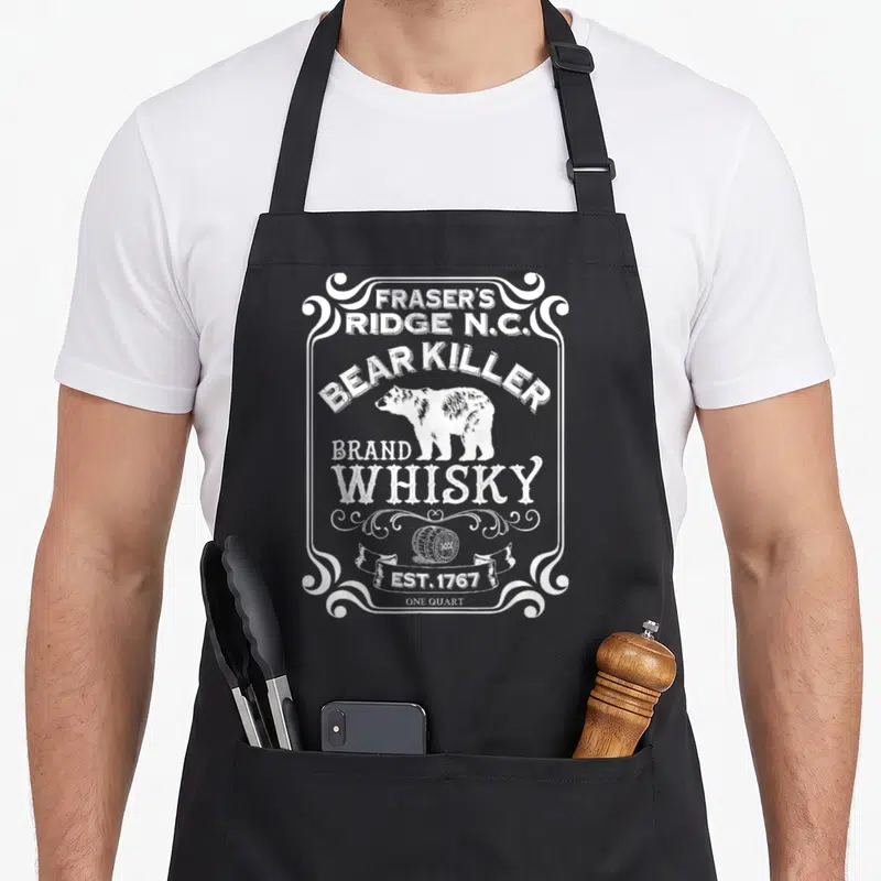 Killer bear apron - White distressed bear label graphic with text Fraser's Ridge Bear Killer Brand Whisky and barrel — shown on a black cotton canvas apron against a white background