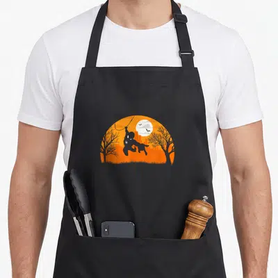 Swing halloween apron - Spider-Man silhouette swinging on web against orange moon and city skyline with two trees — shown on a black cotton canvas apron against a white background