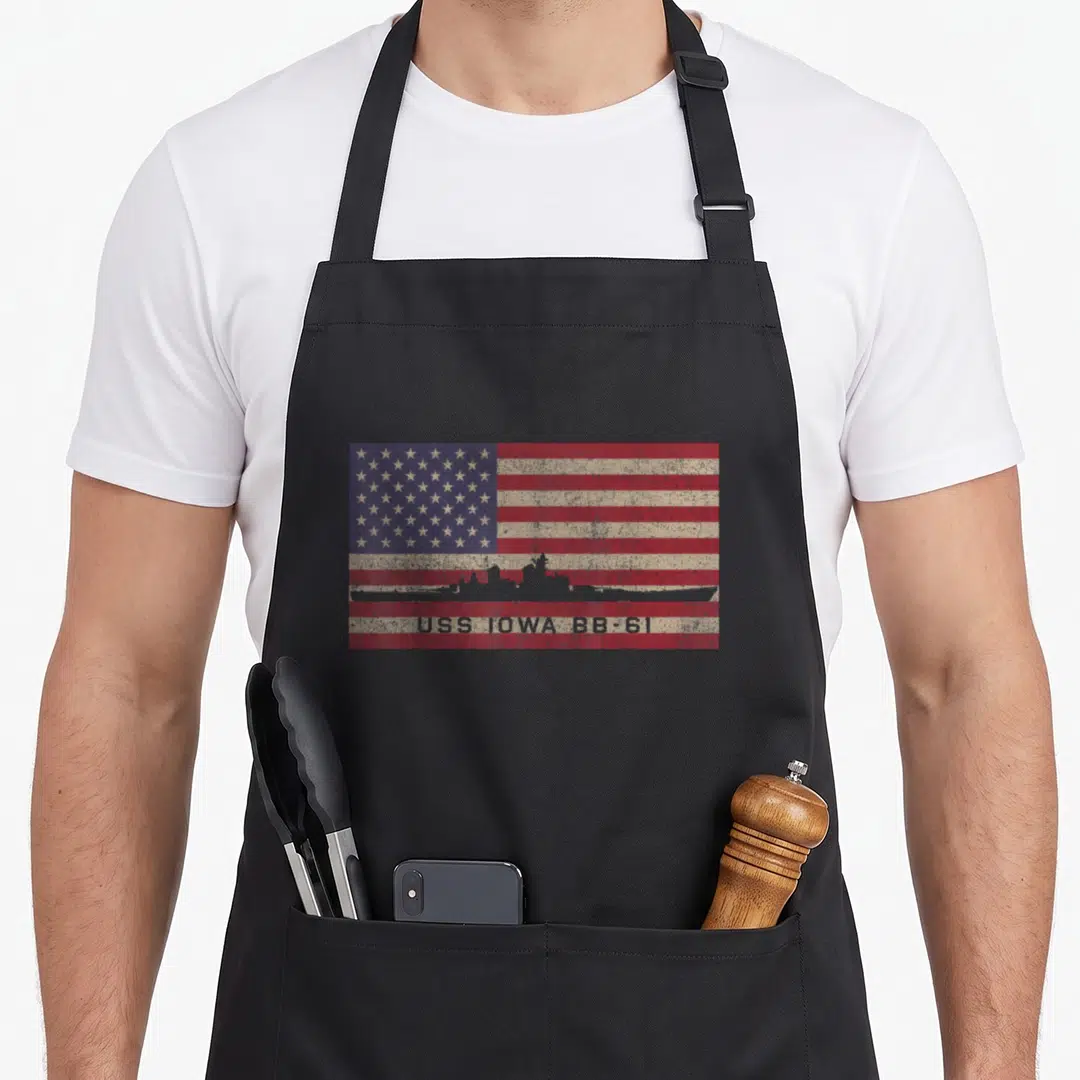 Uss ww2 apron - USS Iowa BB-61 battleship silhouette over distressed American flag with text on bottom stripe — shown on a black cotton canvas apron against a white background