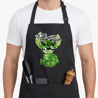 Stitch shenanigan st patrick day aprons - Green Stitch covered in shamrocks with white banner reading LET — shown on a black cotton canvas apron against a white background