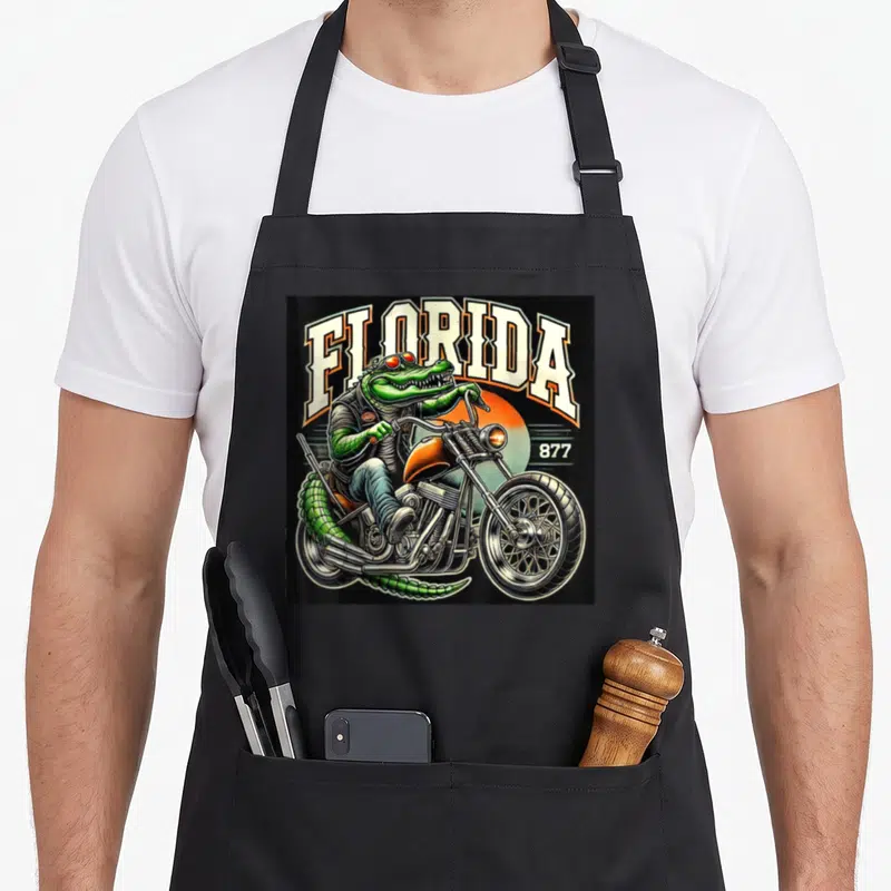 Chopper florida apron - Green alligator riding silver chopper with arched FLORIDA text and orange sun on black — shown on a black cotton canvas apron against a white background
