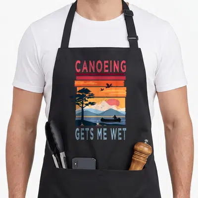 Canoeing wet aprons - Retro sunset landscape with canoeist silhouette and CANOEING GETS ME WET text in red orange — shown on a black cotton canvas apron against a white background