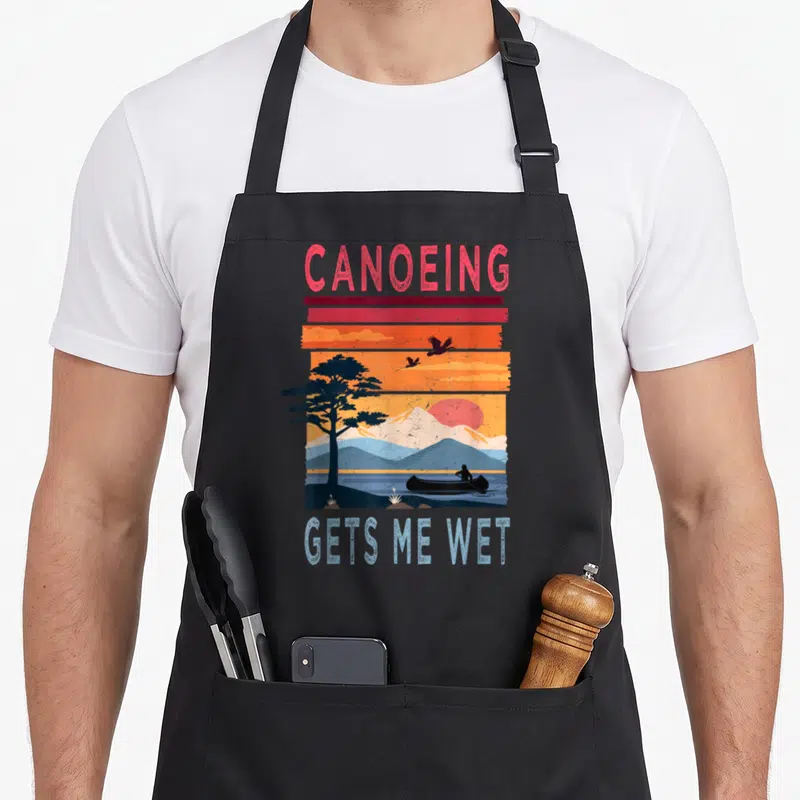 Canoeing wet aprons - Retro sunset landscape with canoeist silhouette and CANOEING GETS ME WET text in red orange — shown on a black cotton canvas apron against a white background