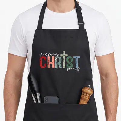 Christian merry christmas apron - Stacked text reading merry CHRIST mas with a cross and multicolored block letters — shown on a black cotton canvas apron against a white background