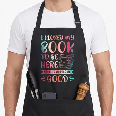 Closed funny spread apron - Watercolor text I closed my book to be here so this better be good next to a stack of books — shown on a black cotton canvas apron against a white background
