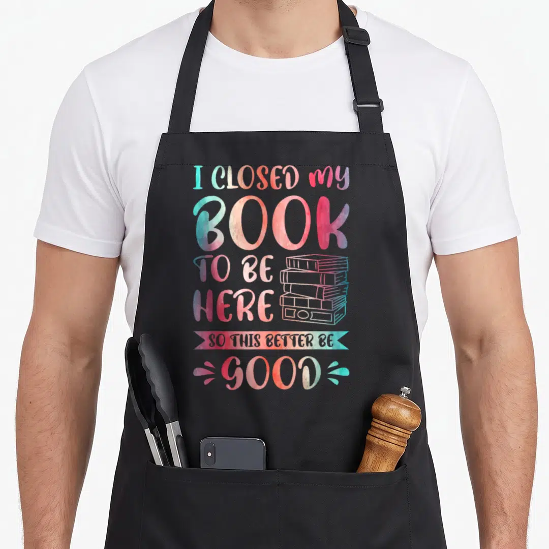 Closed funny spread apron - Watercolor text I closed my book to be here so this better be good next to a stack of books — shown on a black cotton canvas apron against a white background