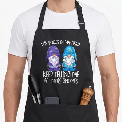 Voice gnome apron - Two cartoon gnomes in purple and blue plaid with white text about voices telling me to get more — shown on a black cotton canvas apron against a white background