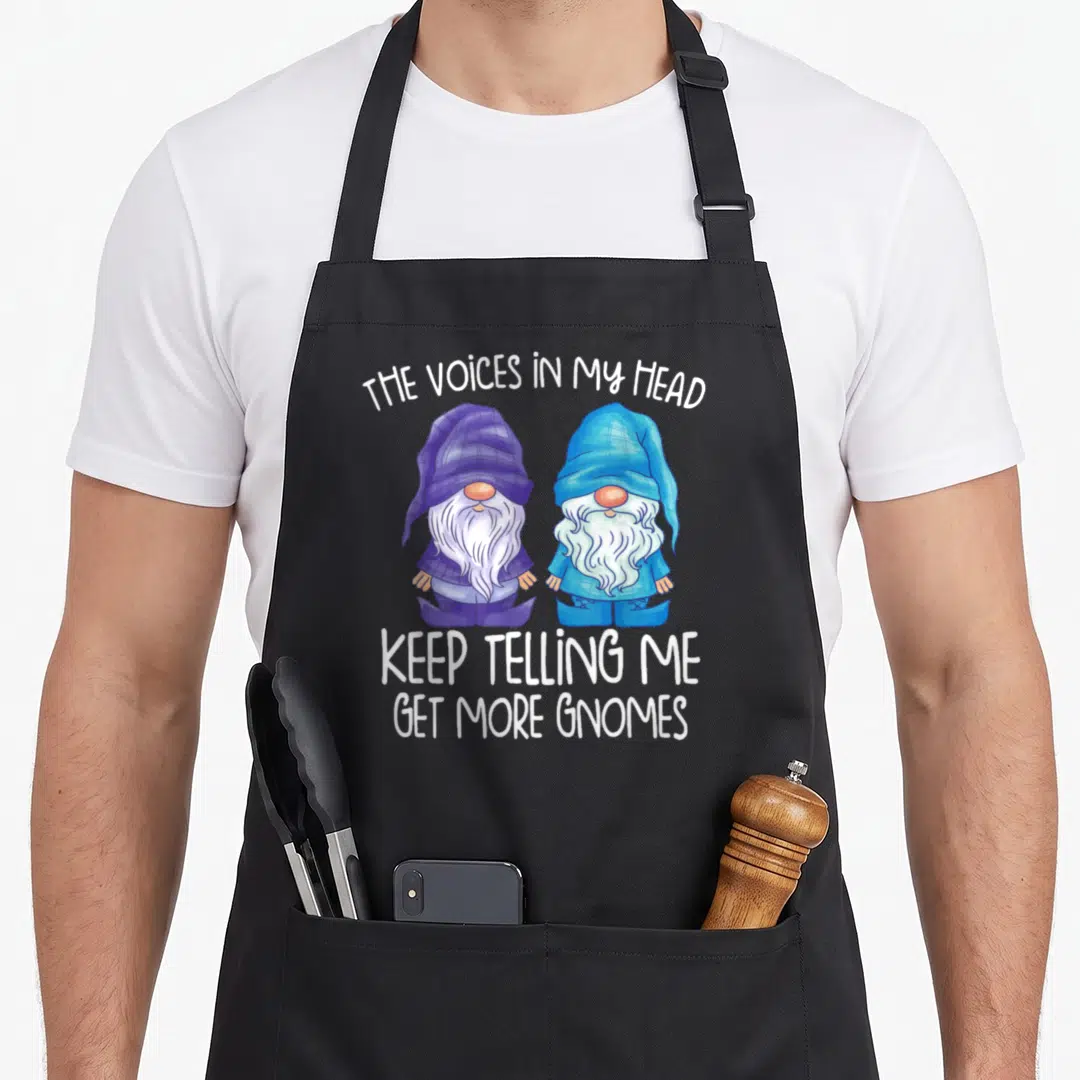 Voice gnome apron - Two cartoon gnomes in purple and blue plaid with white text about voices telling me to get more — shown on a black cotton canvas apron against a white background