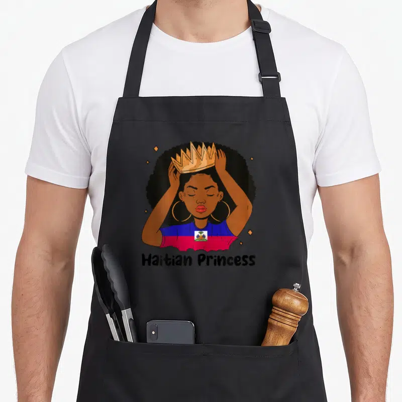 Melanin princess apron - Cartoon woman with gold crown and blue red shirt reading Haitian Princess — shown on a black cotton canvas apron against a white background