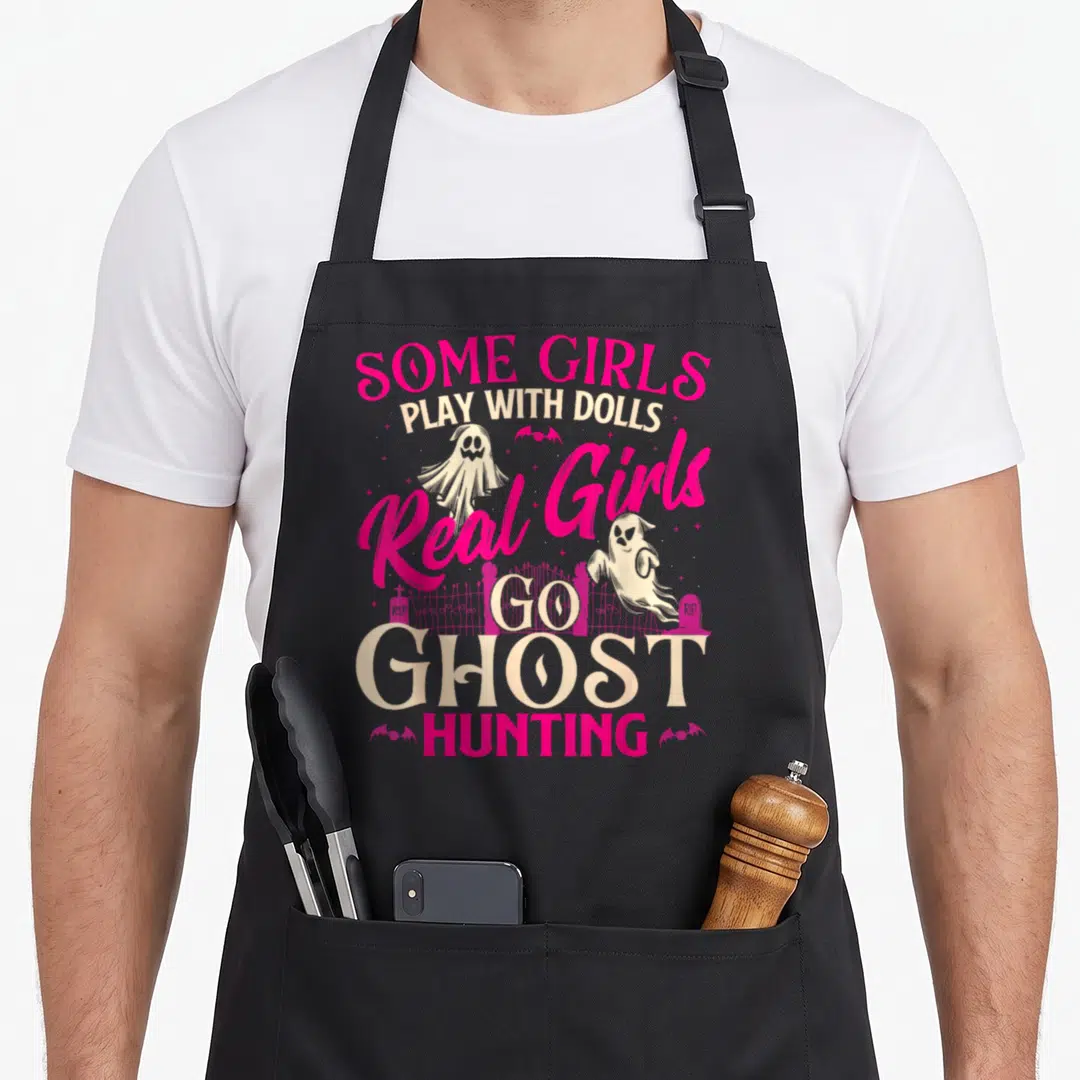 Paranormal ghost apron - Stacked text reading Real Girls Go Ghost Hunting with cartoon ghosts and a tombstone fence — shown on a black cotton canvas apron against a white background