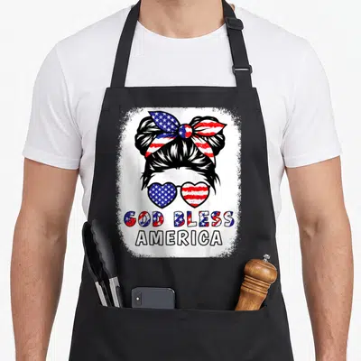 Bless mom daughter apron - Woman bun with flag sunglasses and God Bless America text in distressed red white blue — shown on a black cotton canvas apron against a white background