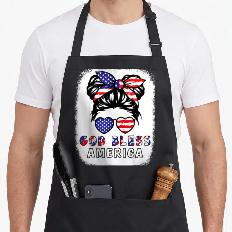 Bless mom daughter apron - Woman bun with flag sunglasses and God Bless America text in distressed red white blue — shown on a black cotton canvas apron against a white background