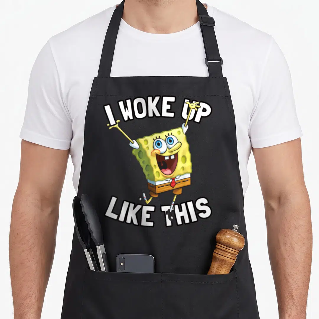 Squarepant woke novelty apron - SpongeBob jumping with I WOKE UP LIKE THIS text in yellow and white — shown on a black cotton canvas apron against a white background