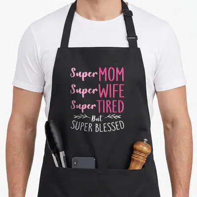 But wife apron - Stacked pink and white text reading Super Mom Super Wife Super Tired But Super Blessed with leaf sprigs — shown on a black cotton canvas apron against a white background