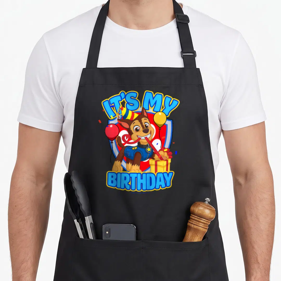 Chase birthday apron - Cartoon brown dog with party hat holding gift box between IT'S MY BIRTHDAY text and balloons — shown on a black cotton canvas apron against a white background