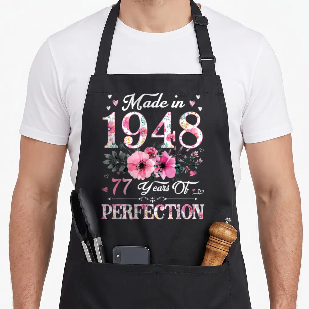 77th vintage floral apron - Watercolor pink flowers and text Made in 1948 77 Years Of Perfection on black apron — shown on a black cotton canvas apron against a white background