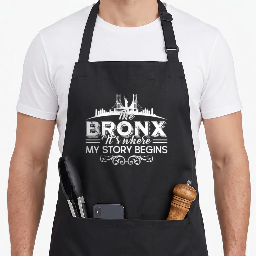 Bronx story of the apron - White distressed skyline with bridge and Statue of Liberty above stacked text reading — shown on a black cotton canvas apron against a white background