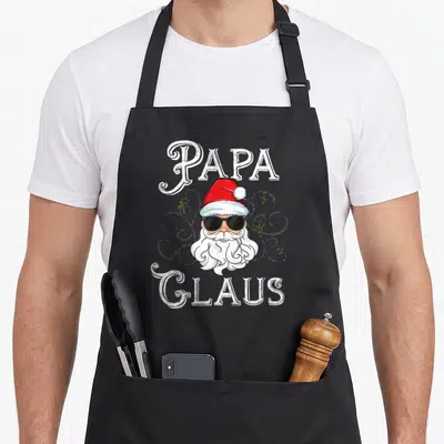 Papa family christmas aprons - Cartoon Santa Claus face with sunglasses surrounded by white swirls and yellow dots — shown on a black cotton canvas apron against a white background