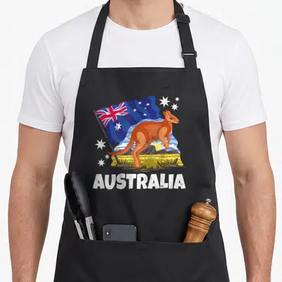 Patriotic kangaroo apron - Cartoon orange kangaroo stands before blue flag and mountains with white AUSTRALIA text below — shown on a black cotton canvas apron against a white background