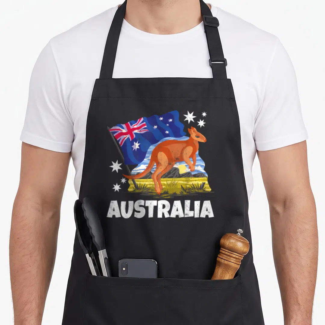 Patriotic kangaroo apron - Cartoon orange kangaroo stands before blue flag and mountains with white AUSTRALIA text below — shown on a black cotton canvas apron against a white background