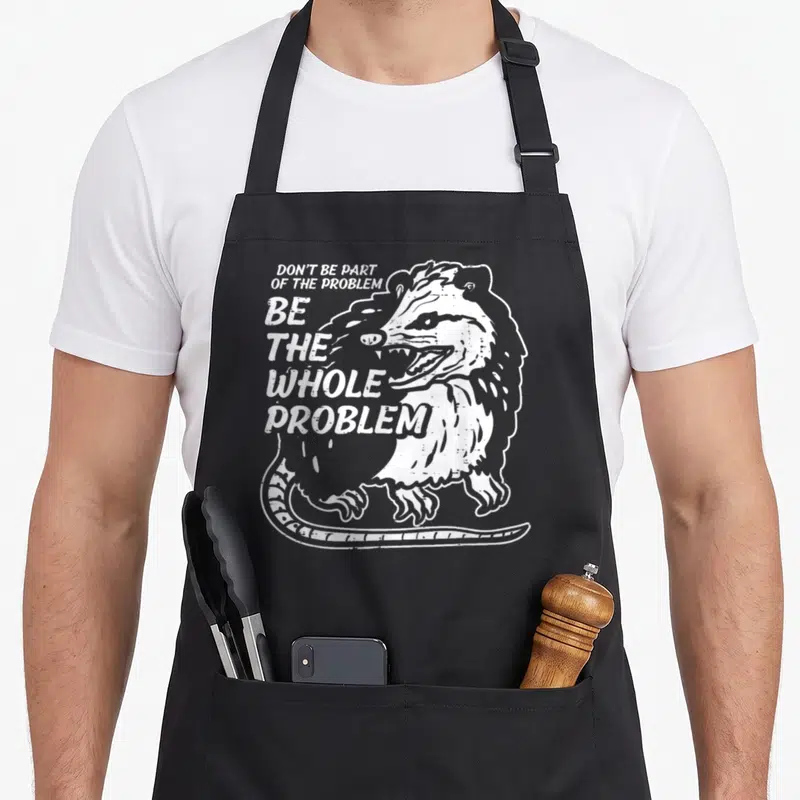 Opossum parts of an apron - White distressed raccoon illustration with stacked text Don't Be Part Of The Problem Be — shown on a black cotton canvas apron against a white background